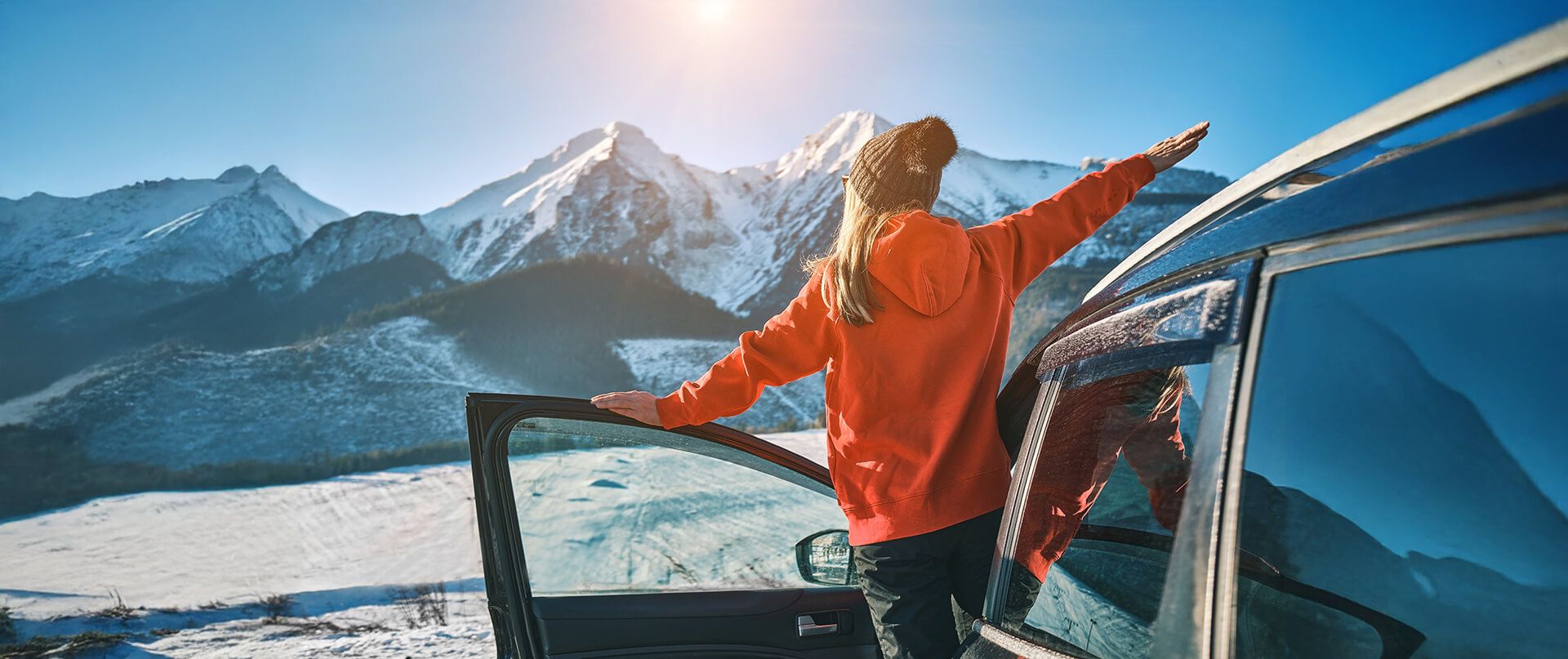 Woman in ski equipment leans out of a van and looks at the snowy mountain panorama.