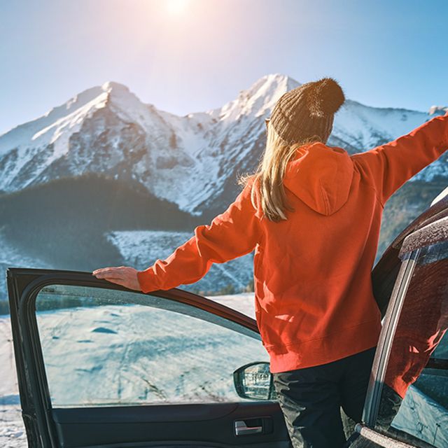 Woman in ski equipment leans out of a van and looks at the snowy mountain panorama.